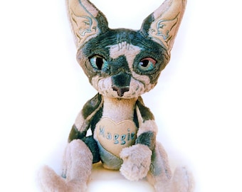 May include: A plush toy resembling a Sphynx cat with grey and beige fur. The cat has large pointed ears with embroidered details, teal-colored eyes, and a heart-shaped patch on its chest featuring the embroidered name Maggie in teal thread.