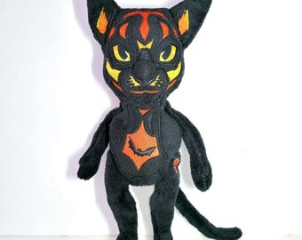 May include: A black plush cat toy featuring embroidered yellow and orange facial markings, large yellow eyes, and pointed ears. The chest area displays an orange diamond-shaped patch with a small black bat silhouette embroidered in the center.