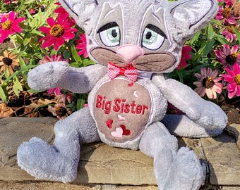 May include: A plush gray cat toy with embroidered features, including large green eyes and a pink nose. It wears a small red and white checkered bow tie. The belly features a tan patch with the embroidered red text Big Sister and small hearts.