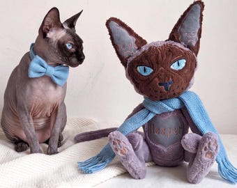 May include: A hairless cat with blue eyes wearing a light blue bow tie sits next to a plush cat toy. The plush toy has brown and purple fur, bright blue eyes, a light blue scarf, and a heart patch on its chest with the name Hesper embroidered.