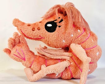 May include: A plush shrimp toy in a soft peach color with a fuzzy texture. The stuffed animal features embroidered black eyes with long lashes, a tuft of peach-colored hair on top, and bright pink stitched details along its segmented body.