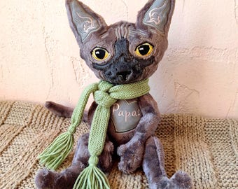 May include: A plush Sphynx cat toy with dark gray fur, large yellow eyes, and pointed ears featuring light gray embroidery. The cat wears a light green knitted scarf with tassels and has a heart-shaped patch on its chest with the word Papas.