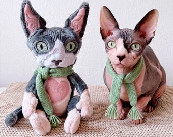 May include: A plush toy cat with gray and white fur, large green eyes, and a light green knit scarf, sitting next to a real hairless Sphynx cat with similar green eyes and an identical light green knit scarf. The plush toy has a pink heart.