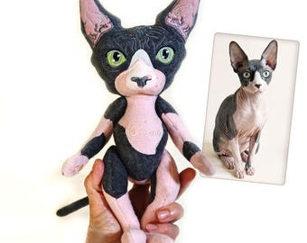 May include: A plush toy modeled after a Sphynx cat with dark gray and light pink fabric patches, large pointed ears, and bright green embroidered eyes. The chest features the stitched name Gizmo, and a reference photo of the cat is included.