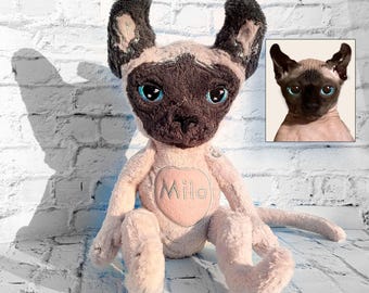 May include: A plush cat toy with light beige fur, dark brown ears, and a dark brown face mask. The toy features bright blue eyes and a pink heart-shaped patch on the chest with the name Milo embroidered in light thread. A photo of a cat is inset.