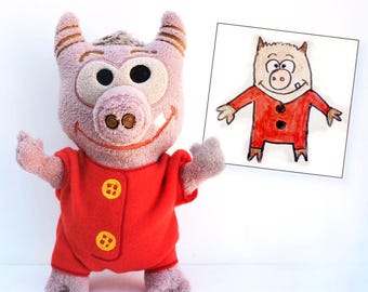 May include: A plush pig character with light pink fur, large eyes, and a single visible tooth, wearing a bright red onesie with two yellow buttons. Beside the plush is a framed drawing of the same character in a red outfit on white paper.