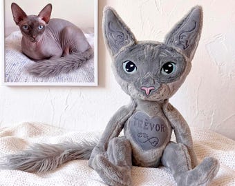 May include: A plush toy cat with grey fur, large pointed ears, and blue eyes. The chest features a heart-shaped patch with the name "TREVOR" and an infinity symbol. A long, fluffy grey tail extends to the left. A framed photo of a cat is visible.