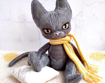 May include: A plush gray cat toy with large amber eyes, a pink nose, and pointed ears. The cat wears a yellow and white striped knit scarf and has the text 'Carmen's Galilo' embroidered on its chest. It sits beside a small white pillow.