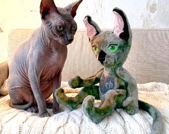 May include: A hairless Sphynx cat with grey skin and green eyes sitting next to a plush toy cat. The plush toy features green fur, large pointed ears with pink inner lining, bright green eyes, and a heart-shaped patch with the text Stitch.