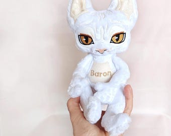 May include: A white plush cat toy with large, pointed ears and amber-colored eyes. The fabric features subtle, raised, swirling patterns across the head and body. A light beige patch on the chest is embroidered with the name Baron in gold thread.