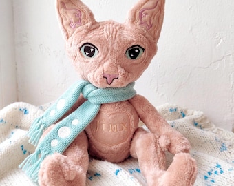 May include: A plush Sphynx cat toy with pink fur, large pointed ears featuring embroidered details, and dark eyes. The cat wears a light blue knit scarf with white polka dots and has a heart-shaped patch on its chest with the name Jinx.
