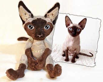 May include: A plush toy resembling a Sphynx cat with light gray and dark brown fur, featuring large ears, blue eyes, and a heart-shaped patch on the chest embroidered with the name Marlo, displayed next to a photograph of a real Sphynx cat.