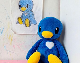 May include: A blue plush toy with a large round head, a yellow beak, and yellow feet. The plush features a white heart patch on its chest and black oval eyes. Behind the plush is a hand-drawn sketch of the same character on white paper.