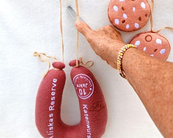 May include: A plush, horseshoe-shaped sausage toy in dark red fabric with white text reading Aliskas Reserve and Katenwurst, featuring a circular 10 Jahre logo. Two round, plush cookie-shaped toys with white and brown spots are attached by twine.