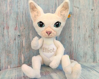 May include: A plush toy resembling a hairless cat with cream-colored fur, large pointed ears, and bright blue eyes. The toy features a white heart-shaped patch on its chest with the name Elliot embroidered in brown thread on the center.