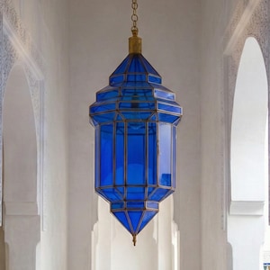 May include: A vibrant blue, geometric lantern hangs from a chain in a white hallway. The lantern's design features a series of glass panels framed by a gold-colored metal. The hallway has arched doorways and a stone floor.