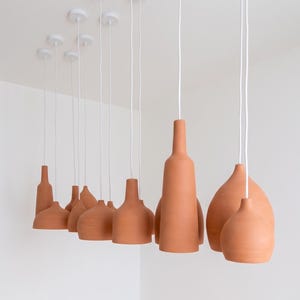 Può includere: Una collezione di lampade a sospensione in terracotta con varie forme a bottiglia e a bulbo, appese a supporti a soffitto bianchi tramite cavi bianchi. Ogni lampada presenta una finitura opaca color arancio terra e una superficie in ceramica liscia.