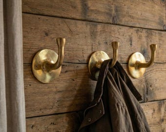 May include: Three brass wall hooks mounted on a rustic wooden wall. One hook holds a brown jacket. The hooks have a circular base and a curved, upward-facing hook. The wood has a natural, aged appearance.