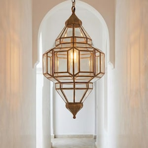 May include: A brass geometric lantern with clear glass panels, suspended from a chain. The lantern houses a warm light bulb, illuminating a white hallway. The design is intricate and decorative.