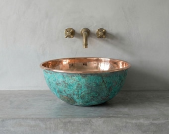 May include: A copper and turquoise patina bowl sink with a polished copper rim. The sink is mounted on a concrete surface, with brass fixtures and a faucet on the wall behind it. The sink has a unique, aged appearance.