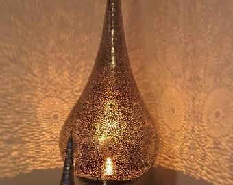 May include: A decorative metal lamp with a teardrop-shaped body and a conical top. The lamp has intricate cut-out patterns that cast a warm, patterned glow on the surrounding wall. A smaller, matching conical lamp is also present.