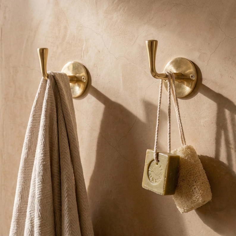 Peut inclure: Deux pat&egrave;res murales en laiton avec une serviette, du savon et une &eacute;ponge v&eacute;g&eacute;tale suspendus. Les crochets ont un design incurv&eacute; et sont mont&eacute;s sur un mur beige. La serviette est beige et le savon est vert.