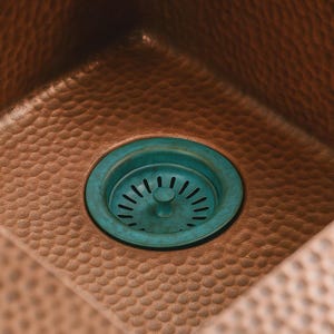 May include: Close-up of a copper sink with a hammered texture. The sink features a teal-colored drain cover with a circular design and radial slots. The copper has a warm, reddish-brown hue.
