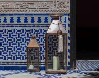 May include: Two antique brass lanterns with glass panels and candles inside. The lanterns are sitting on a white tiled floor with blue and white geometric patterns.
