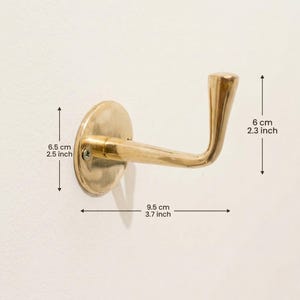 Peut inclure: Un crochet mural en laiton avec un bras incurv&eacute; et une base ronde. Le crochet mesure 9,5 cm de long et 6 cm de haut. La base mesure 6,5 cm de diam&egrave;tre. Le crochet est mont&eacute; sur un mur blanc.
