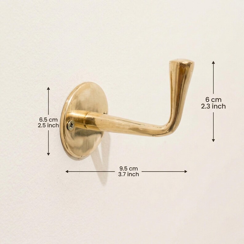 Peut inclure: Un crochet mural en laiton avec un bras incurv&eacute; et une base ronde. Le crochet mesure 9,5 cm de long et 6 cm de haut. La base mesure 6,5 cm de diam&egrave;tre. Le crochet est mont&eacute; sur un mur blanc.
