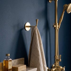Peut inclure: Un crochet &agrave; serviette en laiton avec une serviette beige suspendue, fix&eacute; sur un mur bleu fonc&eacute;. Une pomme de douche et un robinet en laiton sont visibles en arri&egrave;re-plan. Une bouteille d'huile de bain et un savon sont sur une &eacute;tag&egrave;re en dessous.