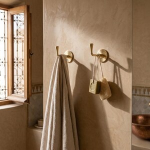Peut inclure: Deux pat&egrave;res murales en laiton, l'une avec une serviette beige, l'autre avec du savon et une &eacute;ponge. Les crochets sont fix&eacute;s sur un mur beige, sugg&eacute;rant une salle de bain. Une fen&ecirc;tre avec un cadre d&eacute;coratif est visible en arri&egrave;re-plan.