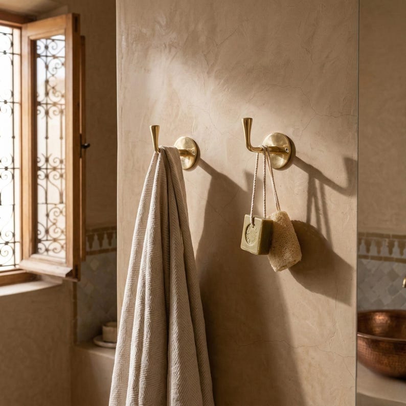 Peut inclure: Deux pat&egrave;res murales en laiton, l'une avec une serviette beige, l'autre avec du savon et une &eacute;ponge. Les crochets sont fix&eacute;s sur un mur beige, sugg&eacute;rant une salle de bain. Une fen&ecirc;tre avec un cadre d&eacute;coratif est visible en arri&egrave;re-plan.