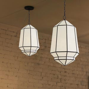 May include: Two white geometric pendant lights with black metal frames hang from the ceiling against a brick wall.