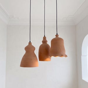 May include: Three terracotta pendant lights hanging from black cords and circular ceiling mounts. Each light fixture features a distinct shape, including a wide base, a tapered neck, and a scalloped edge on the rightmost piece, all in a matte finish.