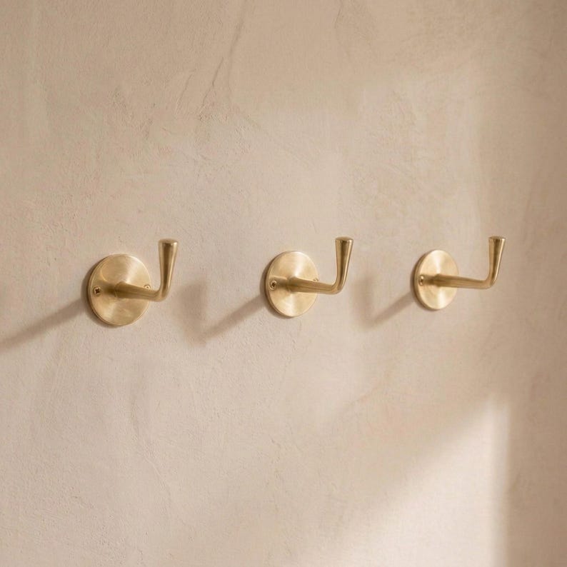 Peut inclure: Trois pat&egrave;res murales en laiton avec une base circulaire et un crochet incurv&eacute;. Les crochets sont mont&eacute;s sur un mur beige clair. Les crochets sont en m&eacute;tal brillant de couleur dor&eacute;e.