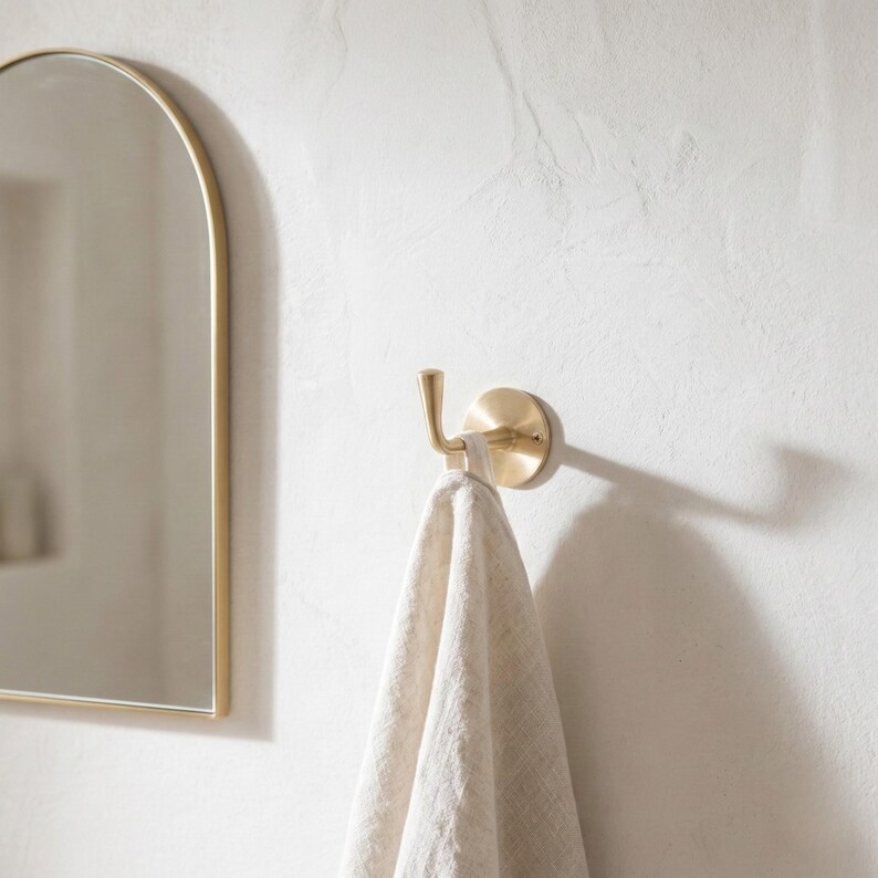 Peut inclure: Un crochet en m&eacute;tal dor&eacute; fix&eacute; sur un mur blanc, tenant une serviette beige. Le crochet a une base circulaire et un bras incurv&eacute;. Un miroir en arc de cercle avec un cadre dor&eacute; est partiellement visible, cr&eacute;ant une esth&eacute;tique minimaliste.