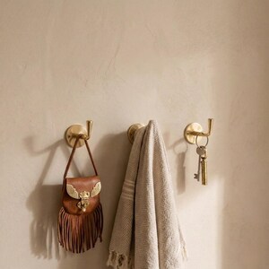 Peut inclure: Trois crochets muraux en laiton avec des objets suspendus. Un petit sac en cuir marron avec des franges est accroch&eacute; au premier crochet. Une serviette beige est sur le deuxi&egrave;me crochet. Des cl&eacute;s sont suspendues au troisi&egrave;me crochet.