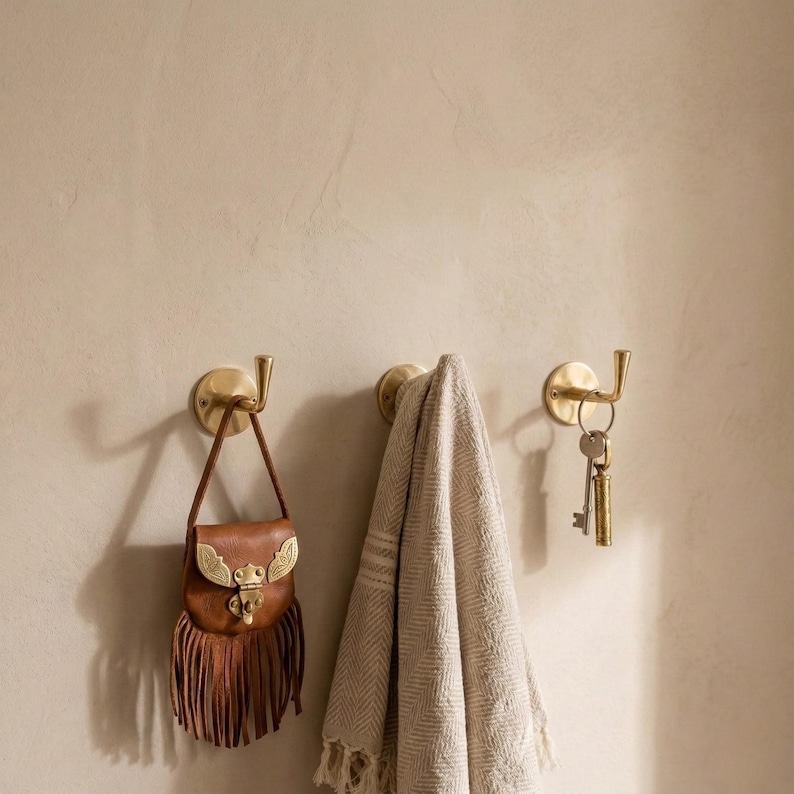 Peut inclure: Trois crochets muraux en laiton avec des objets suspendus. Un petit sac en cuir marron avec des franges est accroch&eacute; au premier crochet. Une serviette beige est sur le deuxi&egrave;me crochet. Des cl&eacute;s sont suspendues au troisi&egrave;me crochet.