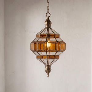 May include: An ornate, hanging pendant light with a geometric design. The light fixture features amber-colored glass panels and clear glass panels, encased in a bronze-colored metal frame. A chain suspends the light from the ceiling. The light is turned on, illuminating the amber glass.