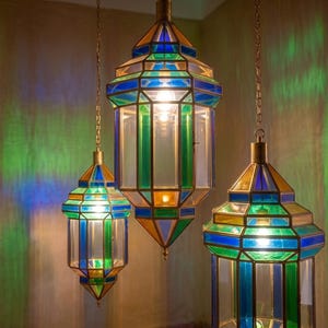 May include: Three hanging pendant lanterns featuring geometric metal frames with clear, blue, and green glass panels. Each lantern is suspended by a thin metal chain and contains an illuminated light bulb, casting colorful light patterns.