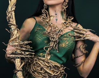 May include: A woman wearing a green dress with a snake-like pattern and gold snake jewelry. She is holding a staff with a snake head at the top. The staff is made of gold and has a snake-like pattern.
