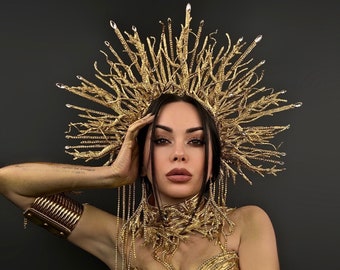 May include: A golden, elaborate headpiece with a sunburst design, paired with a matching gold dress and arm cuffs. The headpiece features crystal accents. The overall aesthetic is regal and theatrical.
