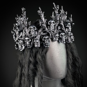 May include: A silver-colored crown featuring numerous skull designs and branch-like structures. The crown is worn on a mannequin head with long, dark dreadlocks. The background is a solid black.