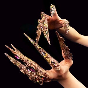 Puede incluir: Adorno de uñas dorado y ornamentado, con intrincados filigranas y piedras preciosas iridiscentes incrustadas. Las largas cubiertas de uñas puntiagudas están diseñadas para usarse en los dedos, creando una estética dramática. Fondo negro.