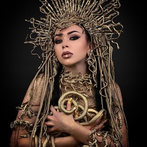 May include: A woman wearing a gold snake crown and a gold snake costume. The snakes are wrapped around her body and head. She is looking at the camera with a serious expression.