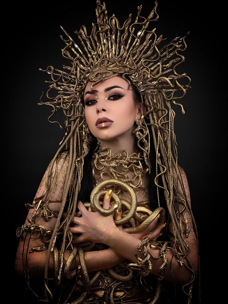 May include: A woman wearing a gold snake crown and a gold snake costume. The snakes are wrapped around her body and head. She is looking at the camera with a serious expression.