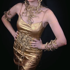 May include: A gold dress with a snake-like design. The dress has a snake-like belt and snake-like bracelets on both arms. The model is wearing gold earrings and has gold makeup on her face.