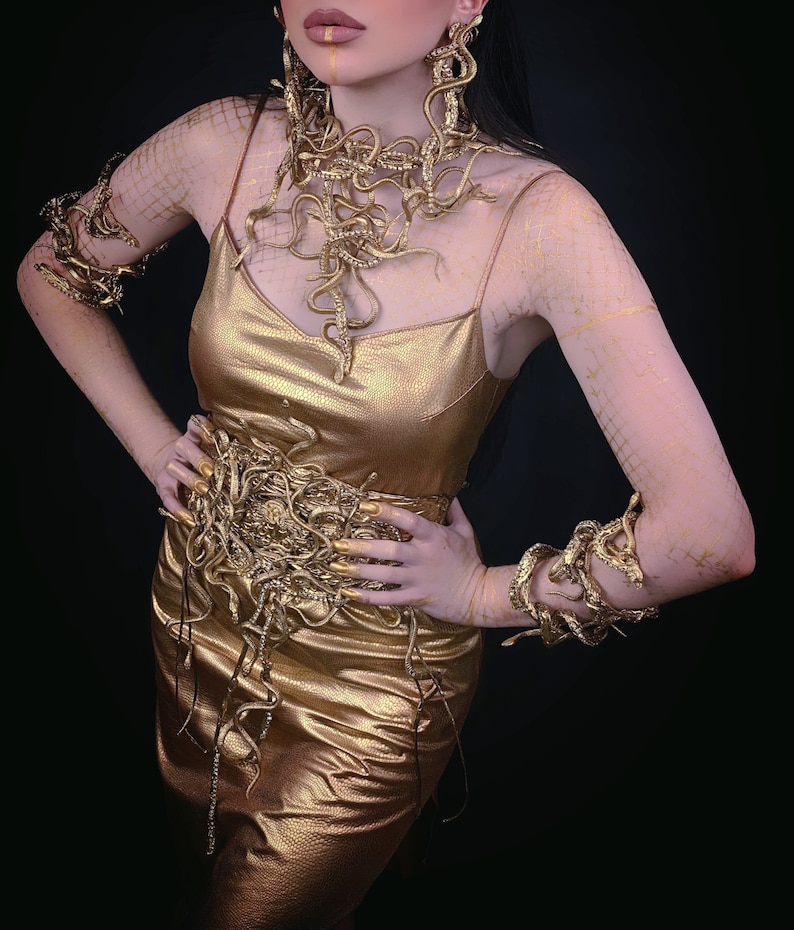 May include: A gold dress with a snake-like design. The dress has a snake-like belt and snake-like bracelets on both arms. The model is wearing gold earrings and has gold makeup on her face.