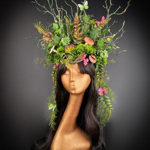 Puede incluir: Una cabeza de maniquí dorada con cabello largo y negro, adornada con una corona de bosque caprichosa. La corona presenta follaje verde, ramas, hongos y mariposas rosas y verdes, creando una estética de cuento de hadas.